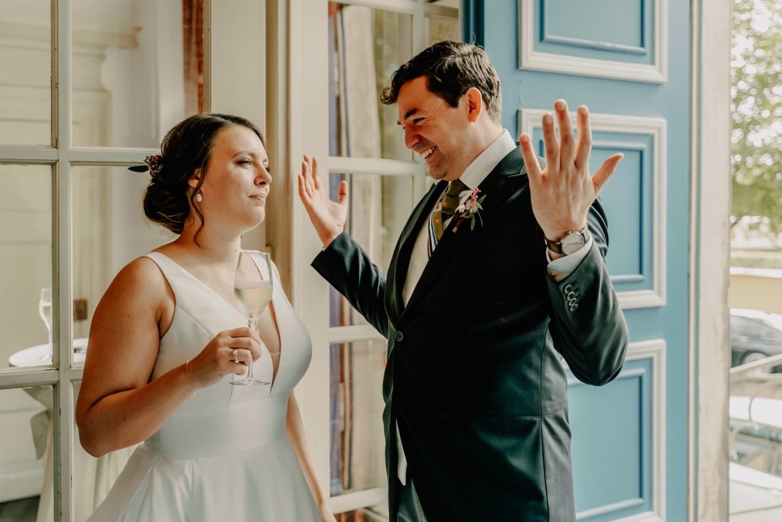 Dieses Bild fängt einen humorvollen und ungezwungenen Moment zwischen dem Brautpaar ein. Die Braut, elegant in einem schlichten weißen Kleid, hält ein Glas Champagner in der Hand und scheint mit einem amüsierten, aber skeptischen Blick auf ihren Bräutigam zu reagieren. Der Bräutigam, gekleidet in einem schicken dunklen Anzug mit einer farbigen Krawatte und Blumenanstecker, gestikuliert lebhaft und strahlt dabei vor Freude. Der Hintergrund mit den großen Fenstern und der blauen Tür verleiht dem Bild eine stilvolle, fast nostalgische Atmosphäre. Dieses Foto zeigt perfekt die Dynamik einer Ehe: liebevolle Momente mit einem Hauch von Humor und Persönlichkeit!