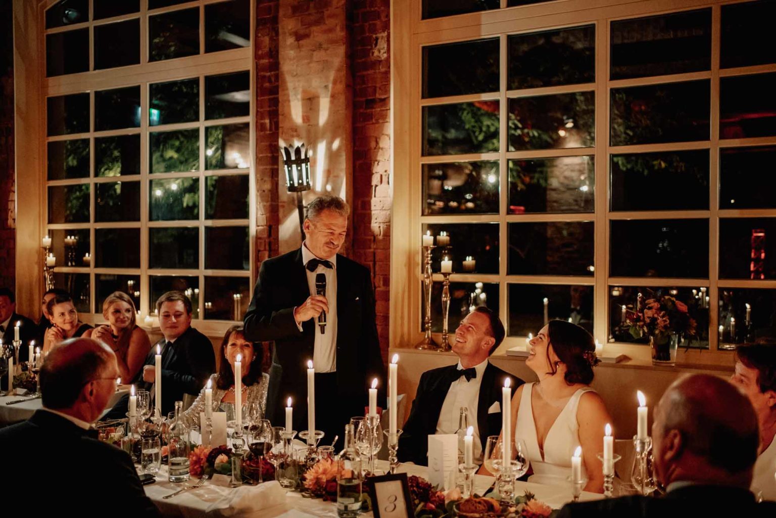 Dieses Bild fängt einen wunderschönen, emotionalen Moment während einer Hochzeitsfeier ein. Ein Mann im eleganten Smoking hält eine Rede und bringt das Brautpaar sowie die Gäste zum Lächeln. Die Stimmung ist warm und festlich, unterstrichen von sanftem Kerzenlicht und einer stilvollen Tischdekoration mit Blumen, Wein und Kerzen. Die großen Fenster und die Backsteinwände verleihen der Szenerie einen charmanten, rustikalen Charakter, während die Reflexionen der Kerzen eine romantische Atmosphäre schaffen. Das glückliche Lächeln des Brautpaars zeigt, wie bedeutungsvoll dieser Moment für sie ist – eine wunderschöne Erinnerung an ihren besonderen Tag.